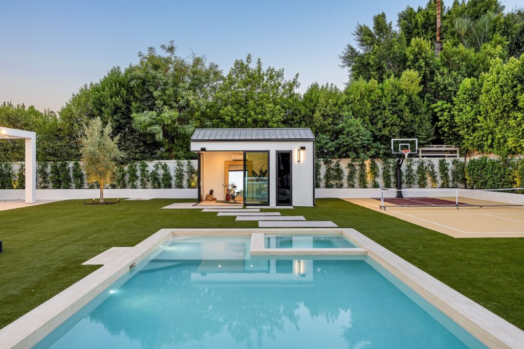 Modern backyard with a pool, basketball court, and garden house surrounded by lush greenery